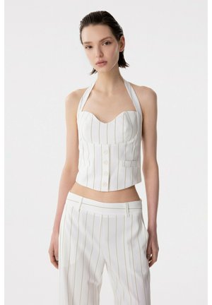 Donna che indossa un top halter bianco a righe sottili con bottoni e pantaloni abbinati a vita alta, in piedi davanti a uno sfondo bianco semplice.