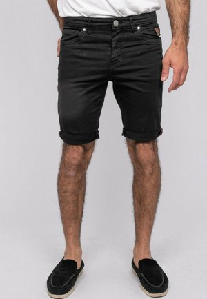 Homme portant un short en denim noir retroussé et des chaussures noires sans lacets, debout devant un fond gris clair uni.
