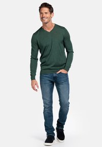 Groene v-hals trui van lichtgewicht breiwerk, gecombineerd met blauwe denim jeans en donkere schoenen. Model staat casual met de handen aan de zijkanten.