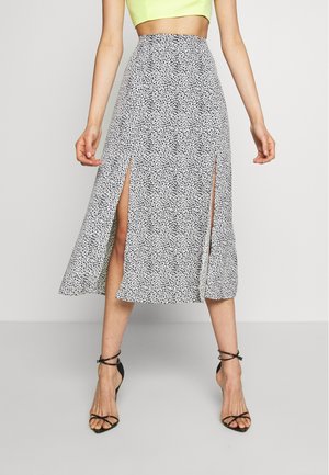 Femme portant une jupe midi blanche à motif noir tacheté avec deux fentes à l'avant, associée à des talons noirs à brides et un top court jaune.