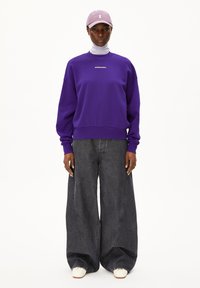 Paarse sweatshirt met ronde hals en logo, gecombineerd met wijde donkerdenim jeans. Witte schoenen en een lichtpaarse pet maken de outfit compleet.