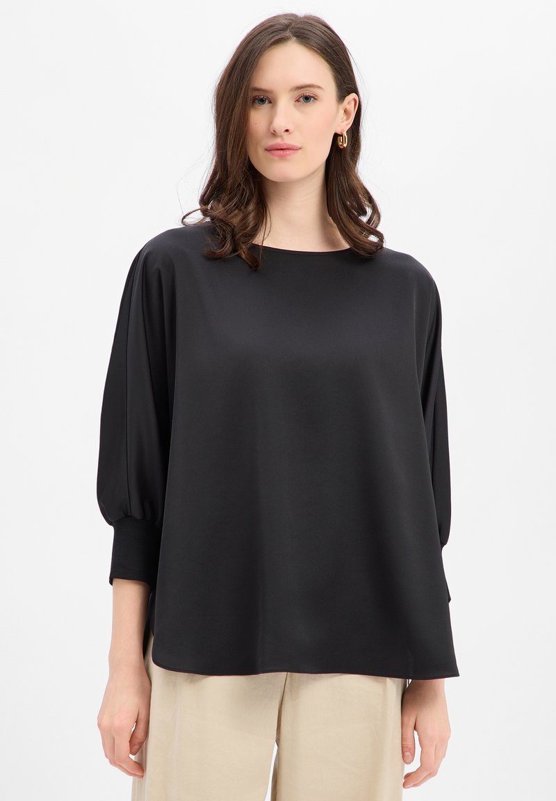 Czarna, luźna bluzka z łodziowym dekoltem i marszczonymi rękawami. Gładka faktura materiału, kontrastowe detale mankietów. Stylizowana z beżowymi spodniami.