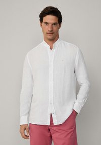 Witte linnen overhemd met lange mouwen, een kraag met knopen en een klein blauw geborduurd detail. Gecombineerd met roze broeken.