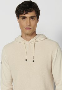 Haut à capuche beige clair en tissu tricoté texturé, featuring un cordon de serrage et des détails noirs sur la manche gauche.