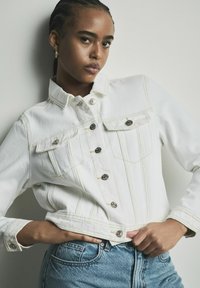Biała kurtka dżinsowa z widocznym przeszyciem, pięcioma srebrnymi guzikami, dwoma kieszeniami na piersi oraz skróconym dołem, zestawiona z jasnoniebieskimi dżinsami.