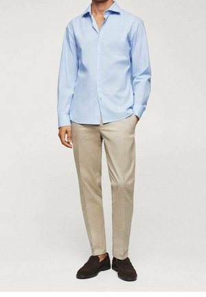 Homme portant une chemise bleu clair à boutons, un pantalon beige et des mocassins marron foncé, debout avec une main dans la poche.