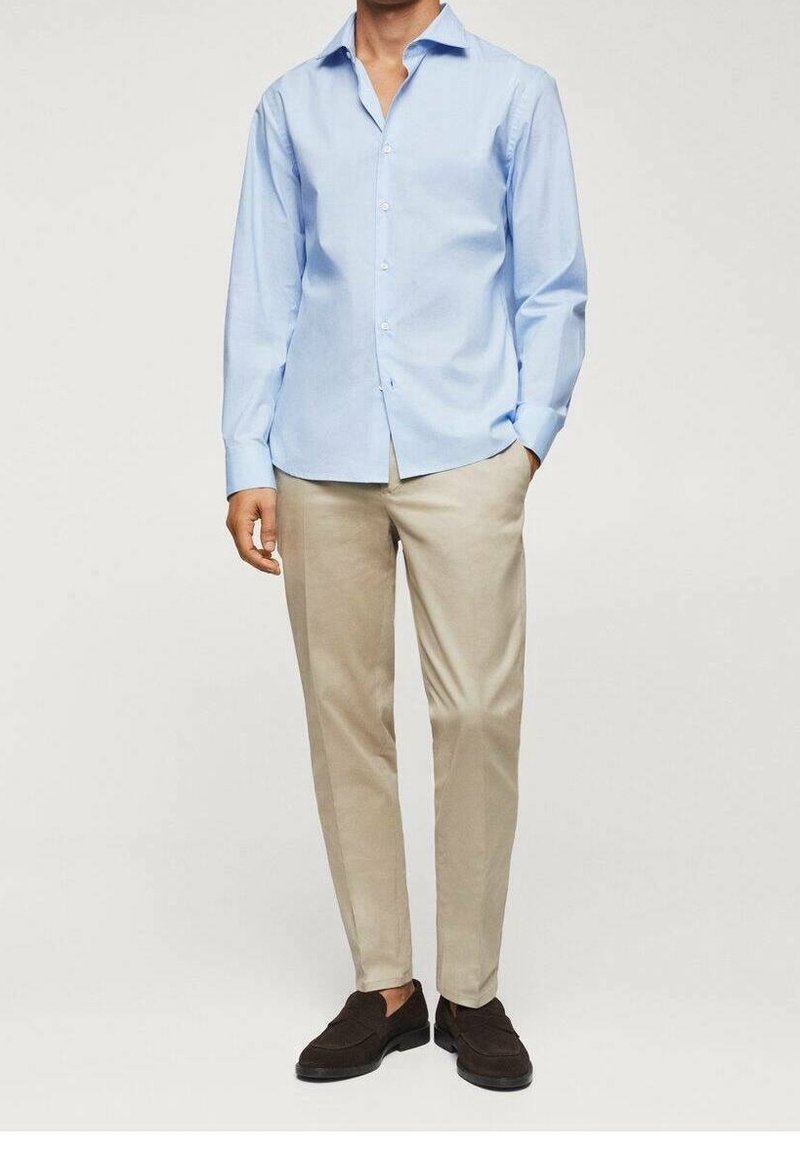 Homme portant une chemise bleu clair à boutons, un pantalon beige et des mocassins marron foncé, debout avec une main dans la poche.