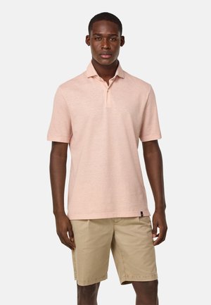 Korte mouwen polo shirt van lichtroze katoen met een drieknops sluiting. Gecombineerd met beige shorts. Model heeft een neutrale uitdrukking.