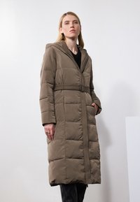 Manteau puffer long et ceinturé en vert olive avec une capuche, texture matelassée, poches latérales et coupe droite, adapté aux temps froids.