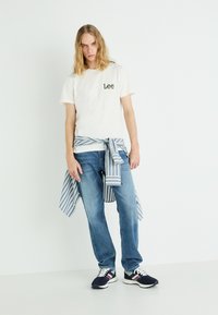 Hvid bomulds T-shirt med grønt "Lee" logo, lyseblå denim jeans og en blå og hvid stribet skjorte bundet i taljen. Sorte sneakers.