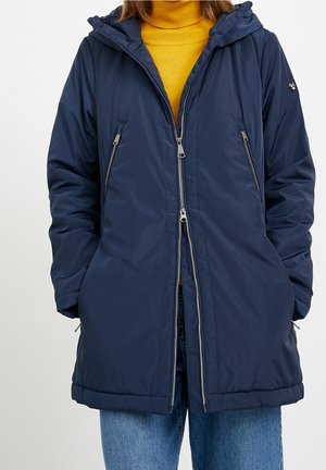 Persoon draagt een marineblauwe winterjas met capuchon en meerdere zilveren ritsen, gecombineerd met een mosterdgele coltrui en blauwe spijkerbroek.