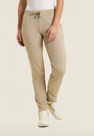 Frau trägt beigefarbene schmale Hose mit Kordelzugbund und weiße Sneaker mit goldenen Akzenten, steht vor hellem Hintergrund.