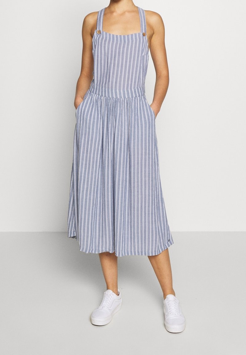 Femme portant une robe midi sans manches à rayures verticales bleues et blanches avec bretelles et des baskets blanches, debout les mains dans les poches.