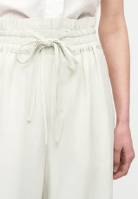 Λευκό παντελόνι με ψηλή μέση, ελαστική ζώνη με ρίγες και κορδόνι στη μέση, φορεμένο με λευκό αμάνικο τοπ και εμφανές δεξί χέρι.