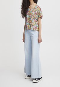 Blouse colorée à motifs avec un col en V, manches courtes bouffantes, associée à un jean large bleu clair et des baskets en toile noires.