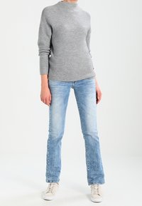 Szary, teksturowany sweter z wysokim kołnierzem, w połączeniu z jasnoniebieskimi spodniami dzwonami i jasnymi trampkami. Na jednolitym tle.