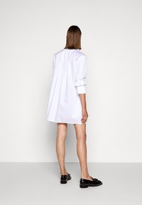 Abito camicia bianco in cotone con maniche a sbuffo e polsini arricciati, caratterizzato da una vestibilità ampia e un orlo dritto. Abbinato a mocassini neri.