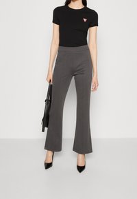 Černé tričko s krátkým rukávem a malým trojúhelníkovým logem, zkombinované se šedými rozšířenými kalhotami. Modelka drží černou kabelku a nosí špičaté černé lodičky.