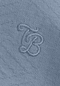 Gebreide stof in lichtblauw met een verhoogd geborduurd monogram "TB" in een decoratief lettertype. Gestructureerde achtergrond met subtiele patronen.