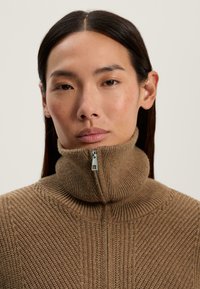 Pulover maro cu guler înalt și fermoar pe jumătate, având o textură cu rippling și un model de tricot vertical. Material moale și confortabil, cu o ajustare strânsă.