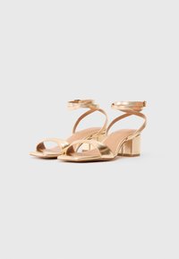 Gold metallic high-heeled sandals with crossed ankle straps, open toe, and block heel, featuring a smooth texture and a simple rectangular shape.