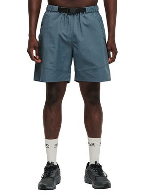 Mann trägt graue knielange Shorts mit integriertem schwarzem Gürtel, weiße Sportsocken mit Text und schwarze Sportschuhe, von der Taille abwärts gezeigt.