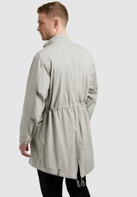 Homme portant une longue veste gris clair avec taille cintrée et fente au dos, debout, légèrement tourné vers la gauche sur un fond uni.