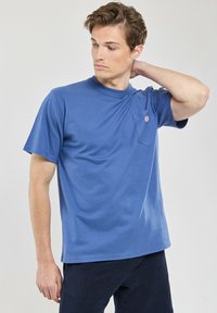 Blaues Baumwoll-T-Shirt mit Rundhalsausschnitt, kurzen Ärmeln und einer linken Brusttasche mit kleinem Logo. Weiche Textur, lässige Passform.