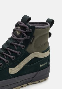 Botas altas verdes y grises con un diseño de ante y textil, presentando un prominente logo de Vans y una marca de Gore-Tex, además de una suela de goma texturizada.