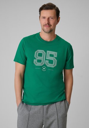 Mężczyzna z krótkimi brązowymi włosami i jasnym zarostem, noszący zielony T-shirt z nadrukiem "95" oraz szare spodnie dresowe, ręce w kieszeniach, neutralne tło.