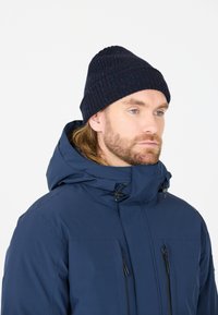 Marineblauwe gebreide beanie met geribbelde textuur en subtiele kleurrijke spikkels, gedragen met een bijpassende blauwe waterdichte jas met ritsen.