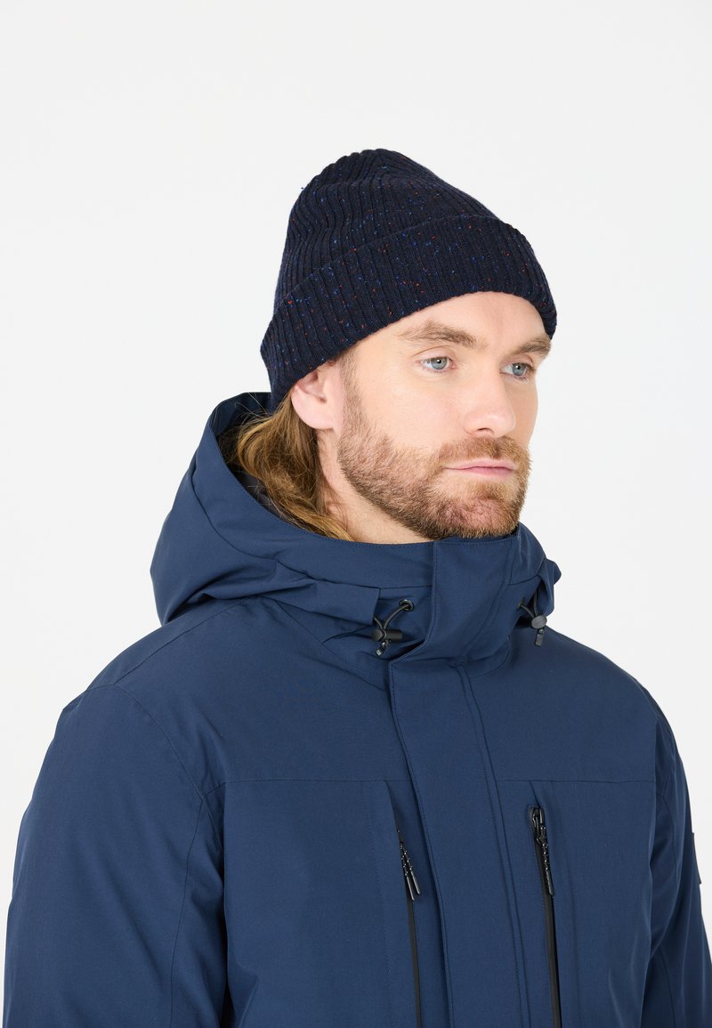 Marineblauwe gebreide beanie met geribbelde textuur en subtiele kleurrijke spikkels, gedragen met een bijpassende blauwe waterdichte jas met ritsen.
