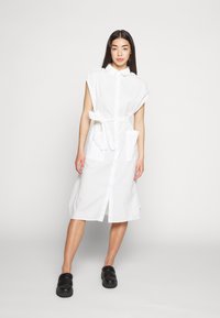 Vestido de camisa branco riscado com mangas curtas, com botões na frente, cintura cintada e dois bolsos frontais. Usado com tamancos pretos.