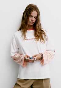 Weißes, übergroßes Sweatshirt mit orange gestreiften Ärmeln und einem passenden Halsausschnitt. Der Text "ICH BRAUCHE EINEN EISBECHER" in Braun. Das Modell hält einen blauen Gegenstand.