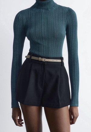 Petrolfarvet ribstrikket rullekravesweater kombineret med højtaljede sorte shorts og et lyst beige bælte på en stående person.