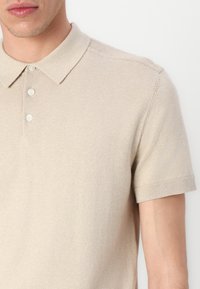 Bež majica polo kratkih rukava izrađena od mekanog pletenog materijala. Ima klasičnu kragnu, placket s tri dugmeta i rebraste rubove rukava.