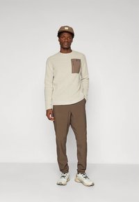 Svetlo bež teksturiran pulover z rjavim žepom na prsih, kombiniran z rjavimi hlačami in sivimi supergami. Priložnostni slog s preprosto kapo.