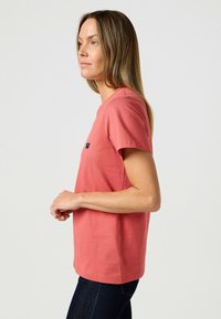Korallenfarbener Baumwoll-T-Shirt mit kurzen Ärmeln, Rundhals-Ausschnitt und einem kleinen Logodetail auf der Brust. Kombiniert mit dunkelblauen Jeans.