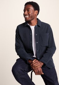 Navy corduroy jacket with a button front, chest pocket, and textured finish, worn over a white t-shirt and paired with dark denim jeans.