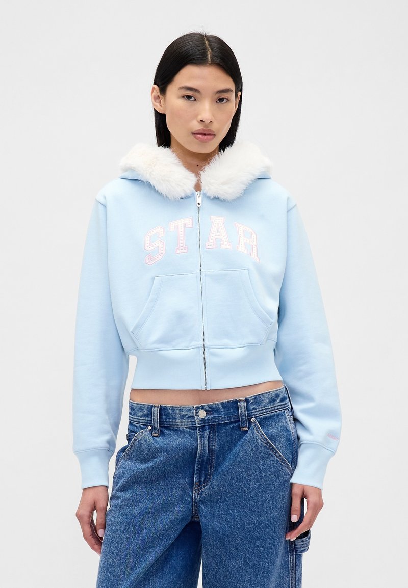 Felpa corta azzurro chiaro con colletto in ecopelliccia bianca, zip frontale e scritta "STAR" in rosa. Presenta due tasche laterali e polsini a costine.