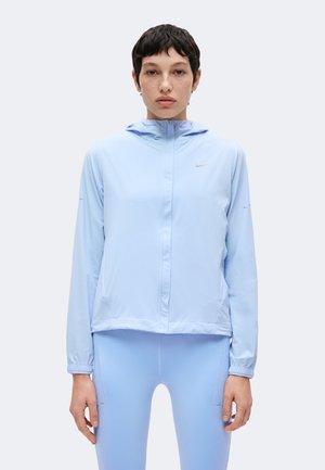 Person, die frontal steht und eine hellblaue Kapuzenjacke sowie passende Leggings auf weißem einfarbigem Hintergrund trägt.