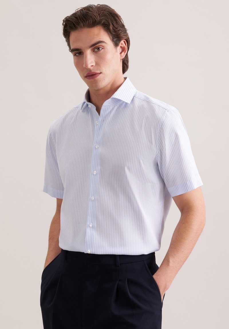 Jonge man met een lichtblauw, dun gestreept overhemd met korte mouwen en knopen, gecombineerd met een donkere broek, handen in de zakken, neutrale achtergrond.