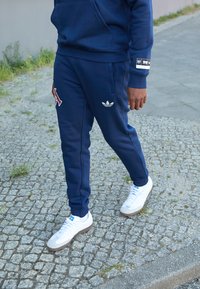 Temno modra trenirka z rdečim logotipom "A" in belim Adidas trifoilom. Bele superge z sivo-poudarjenimi detajli. Gladka tkanina, oblikovanje v ožjem kroju.