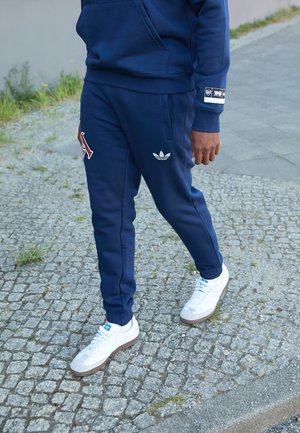 Marineblå træningsdragt med rød "A" logo og hvid Adidas trefoil. Hvide sneakers med grå accenter. Glat stof, slim fit design.