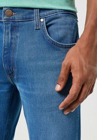 Jeans in denim blu medio con taglio classico, dotati di cuciture arancioni, bottone metallico e dettaglio sulla tasca anteriore. Mano appoggiata sul tessuto.