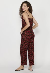 Zwarte bloemen-jumpsuit met rode en groene patronen, voorzien van dunne spaghettibandjes, een taillering en zijzakken. Textuurstof voor comfort.