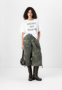 Witte oversized t-shirt met de tekst "SAVAGE NOT AVERAGE", gecombineerd met groene camouflage cargo-broeken en kniehoge bruine leren laarzen.