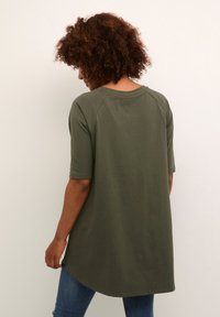 Tunique vert olive, longueur genoux, avec un col rond et des manches raglan courtes. Texture douce, design décontracté et ourlet incurvé. Jeans en denim visibles.