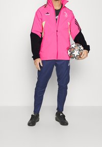 Lyserød og sort træningsjakke med lynlås, der har et Juventus logo, parret med marineblå bukser og sorte fodboldstøvler, mens der holdes en mønstret fodbold.