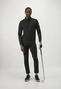 Juodas zip-up ilgos rankovės marškinys, juodos kelnės, baltas golfas, juodi sportbačiai su baltais padais, laiko golfą, stovi ant vientiso fono.